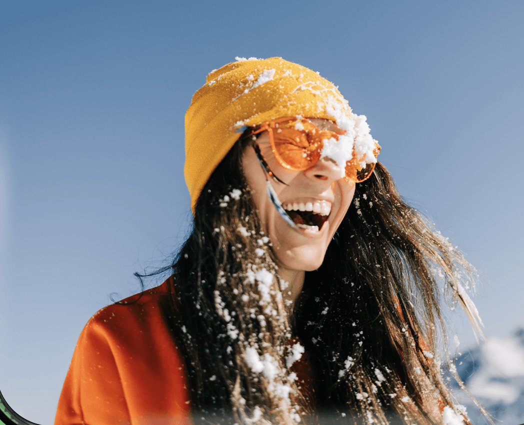 Laughing woman with snow-covered yellow beanie and orange sunglasses wearing red jacket outdoors against clear blue winter sky, joyful ski holiday lifestyle portrait