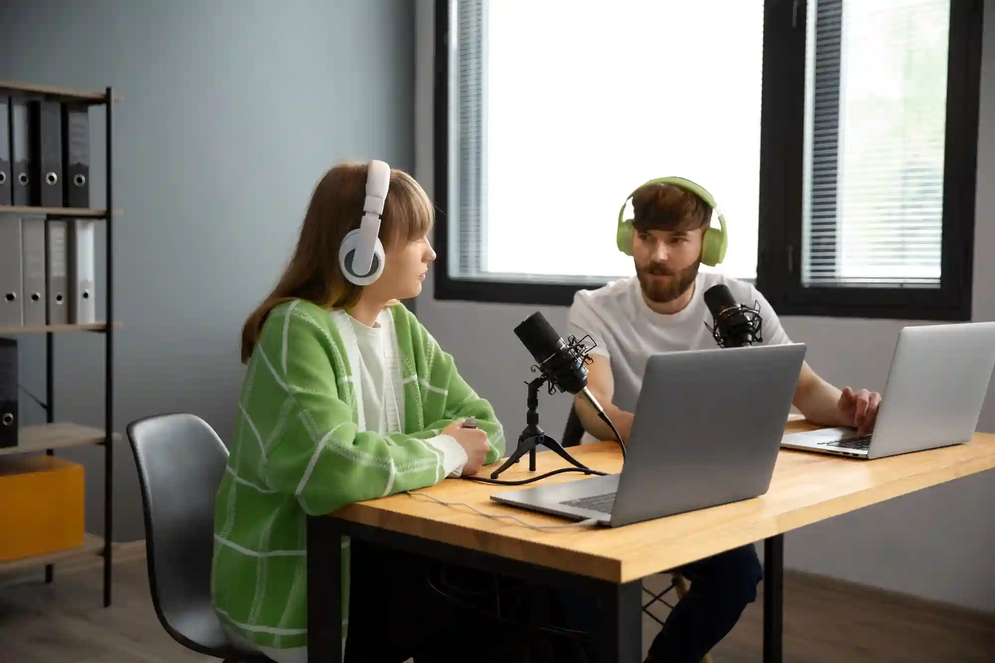 A man and a woman wearing headphones are recording a podcast using microphones and laptops at a desk.