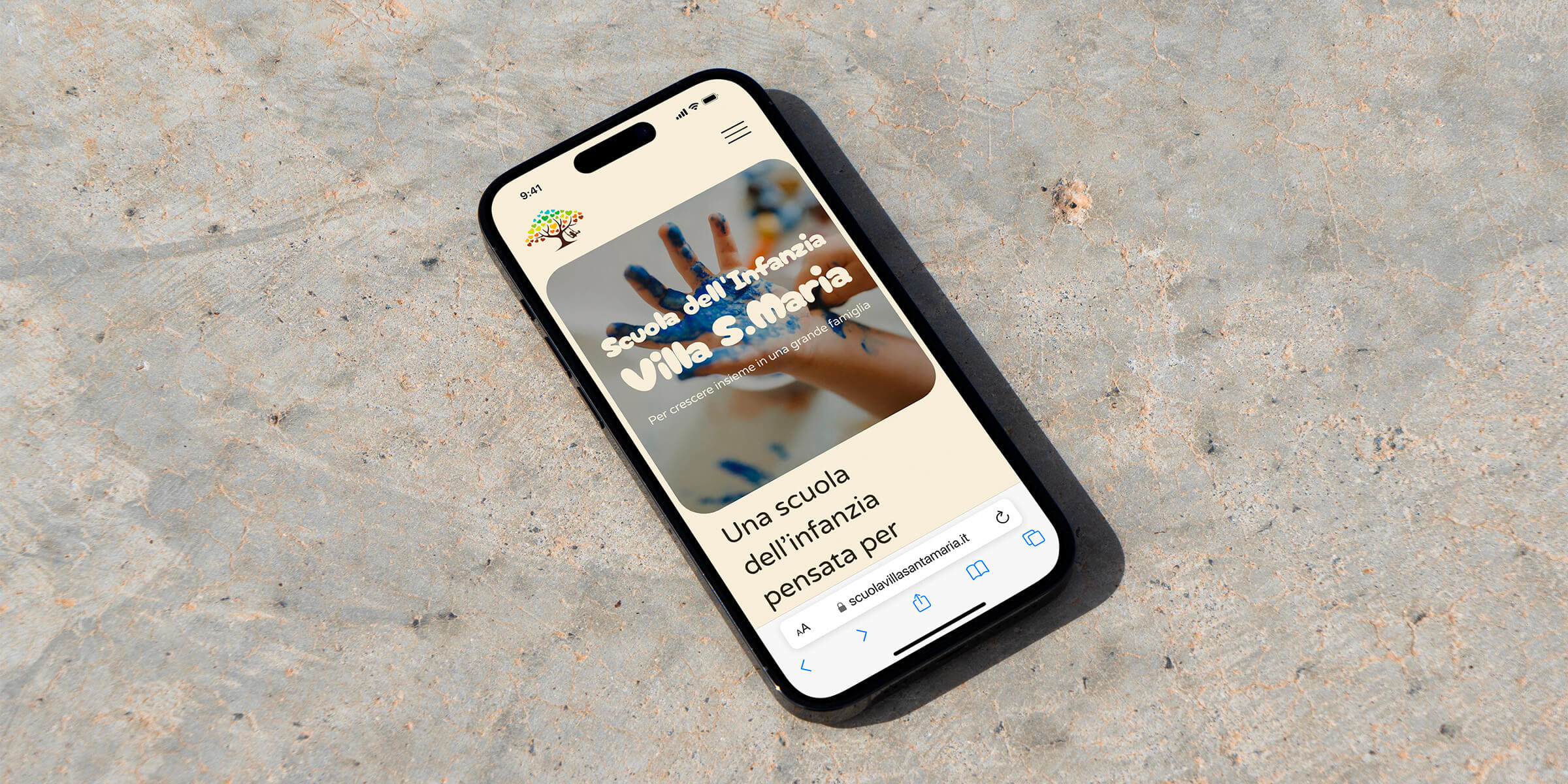 A smartphone displaying a nursery website of Villa S. Maria with colourful handprints and playful text lies on a textured, sandy surface.