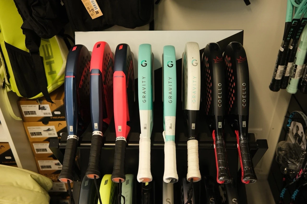 Verschiedene Padel-Schläger von HEAD, darunter Modelle der Gravity- und Coello-Serie, ordentlich in einem Regal präsentiert. Die Schläger haben unterschiedliche Farben, darunter Blau, Pink, Weiß und Schwarz. Im Hintergrund sind Tennistaschen, Schuhkartons und weitere Schläger sichtbar.
