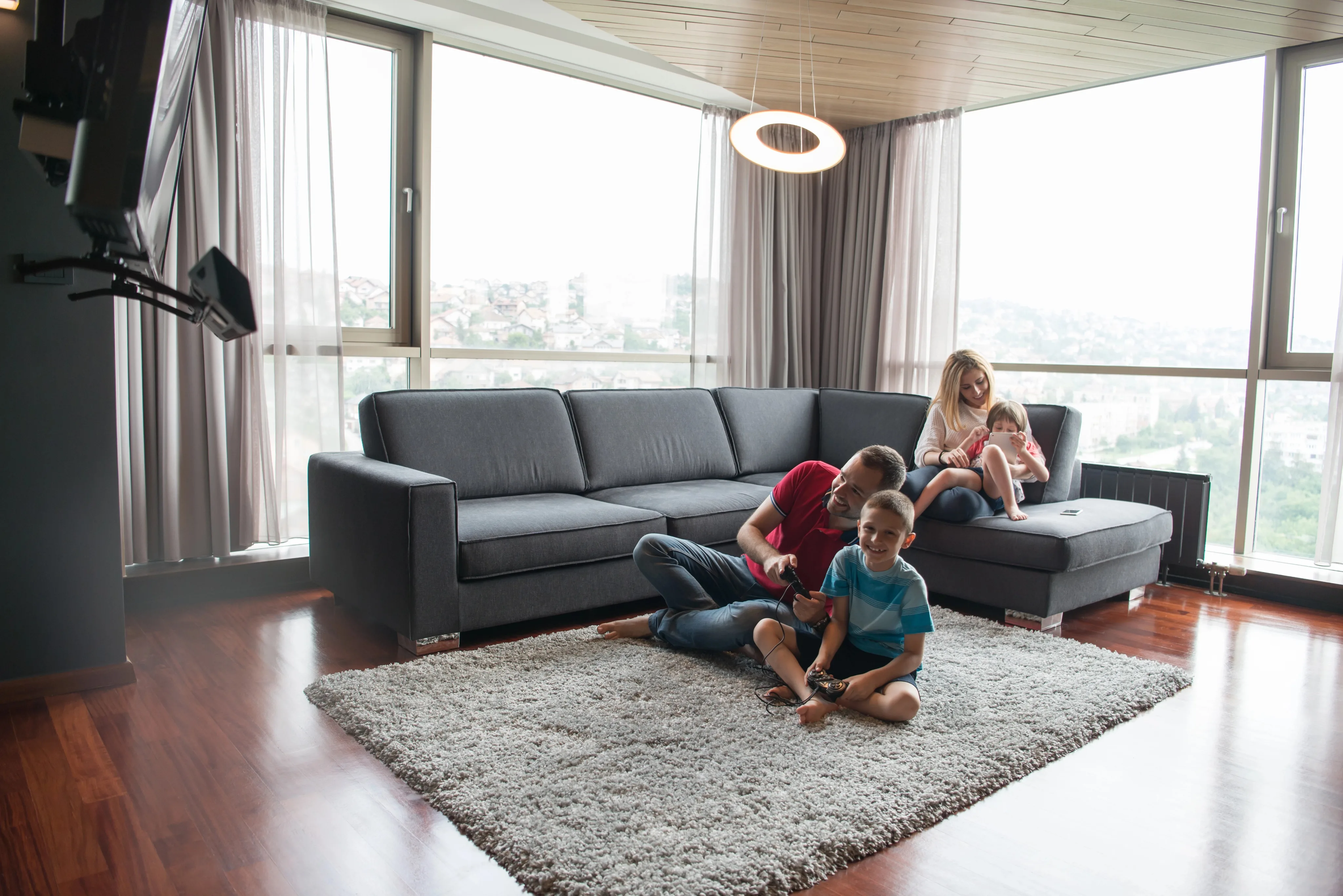 A family of four relaxes in a modern high-rise living room with a gray sectional sofa, gray area rug, and floor-to-ceiling windows overlooking a cityscape.