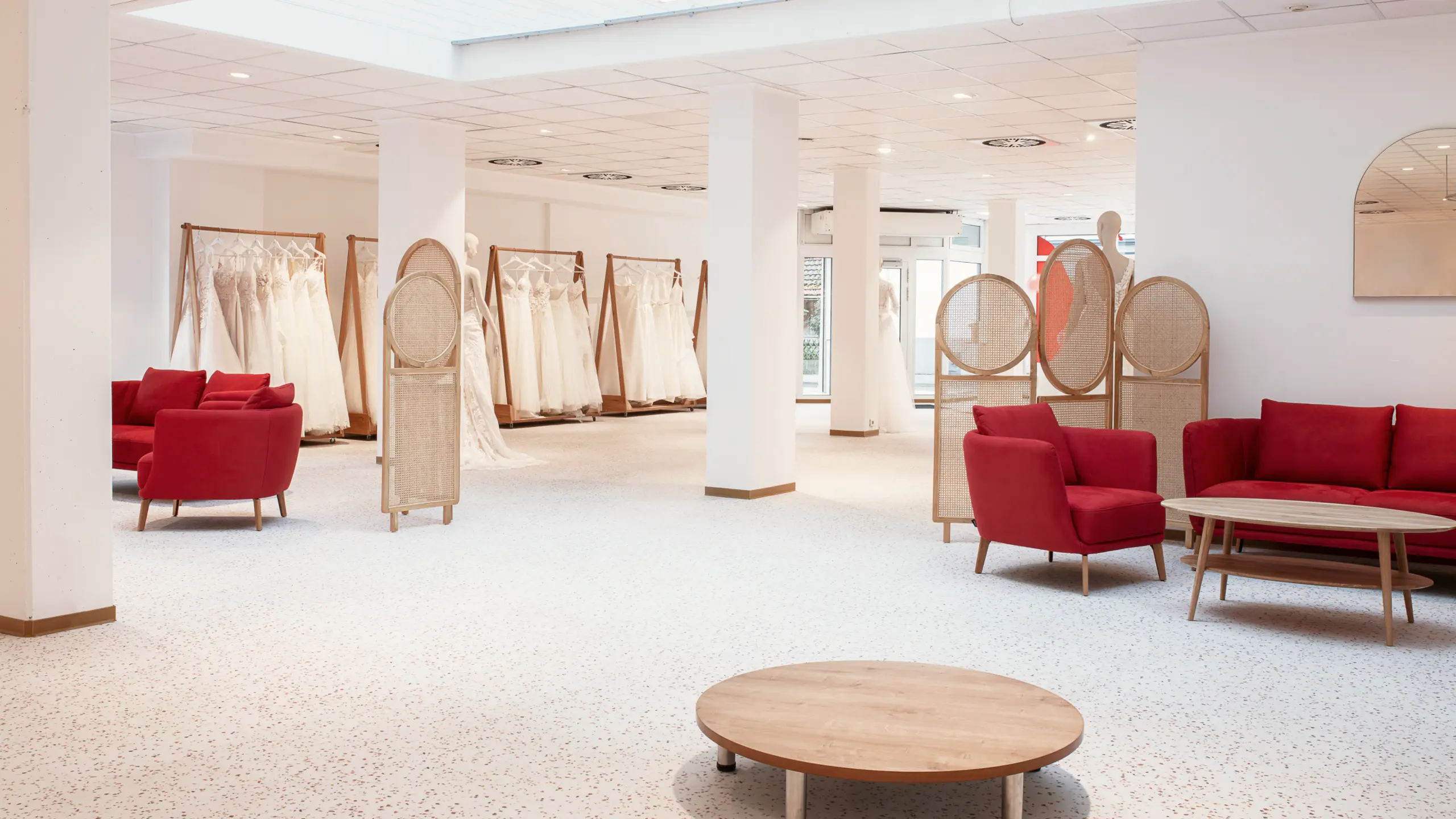 Blick in die helle Boutique von Kerst Hochzeitsmode in Ludwigsburg mit Brautkleidern an Holzstangen, roten Samtmöbeln, Terrazzo-Boden und Rattan-Raumteilern.