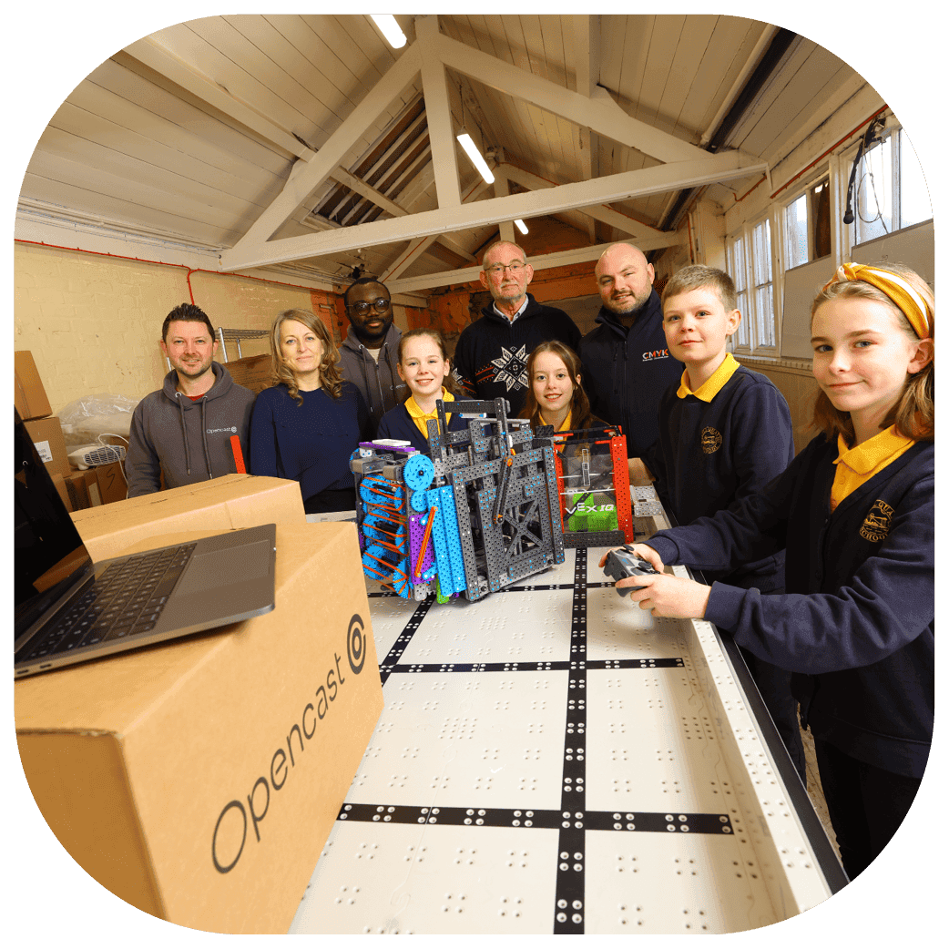 Group of students and adults gathered around a robotics table with assembled mechanical parts and a laptop.