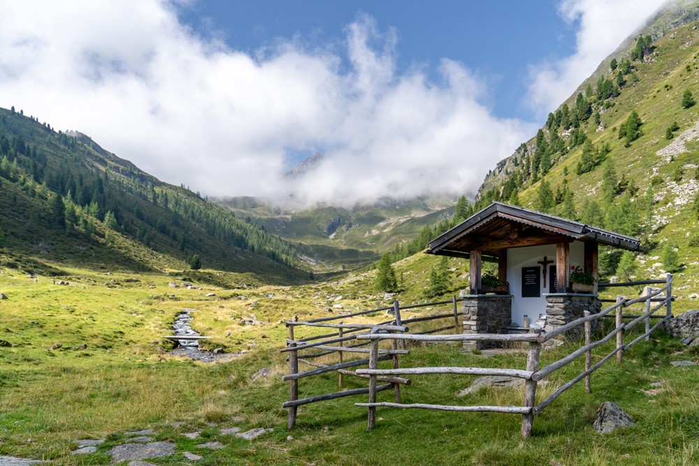 Bergkapelle, umgeben von grünem Gras und majestätischer, wolkiger Berglandschaft in der Natur.