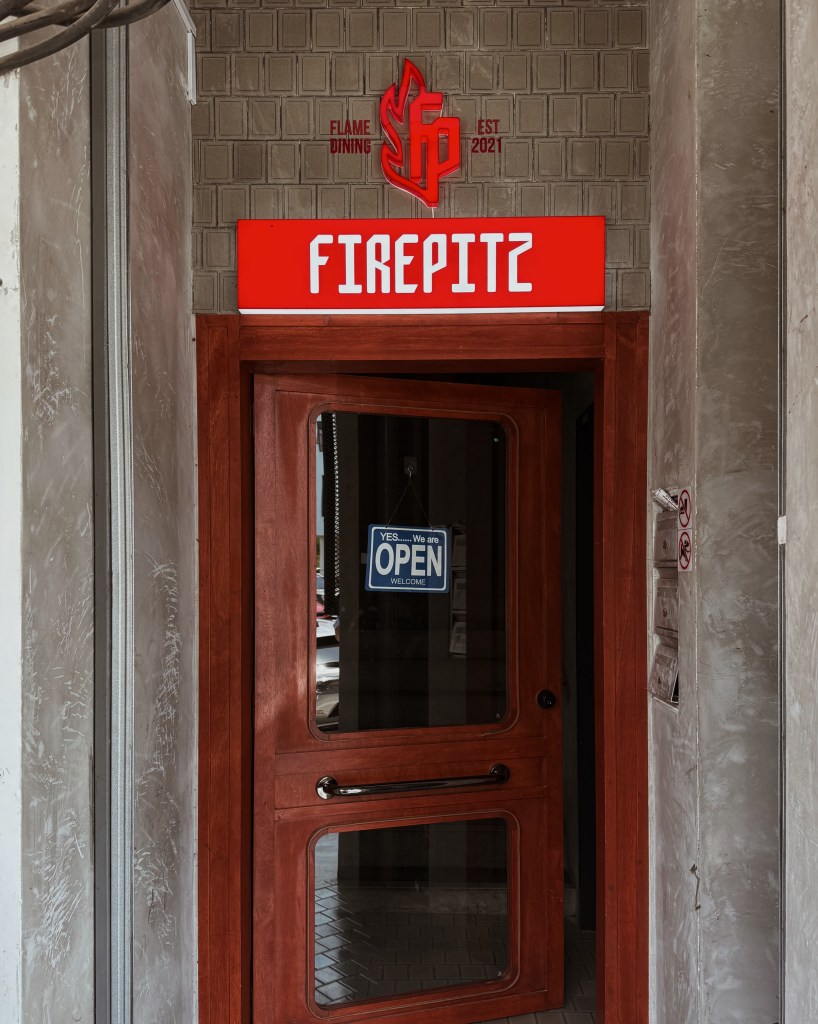 Entrance of Fire Pitz steakhouse featuring a wooden door with an 'OPEN' sign, showcasing the restaurant's logo and name above.