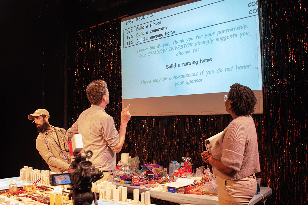 Two performers look at a projection screen showing voting results. Another performer peers at the audience.