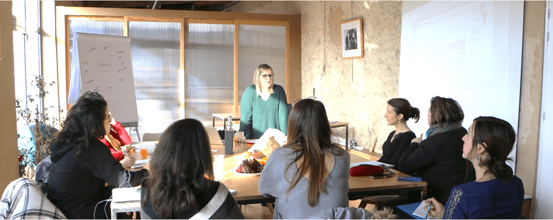 Groupe de participantes assises autour d'une table en formation, une formatrice debout face à elles