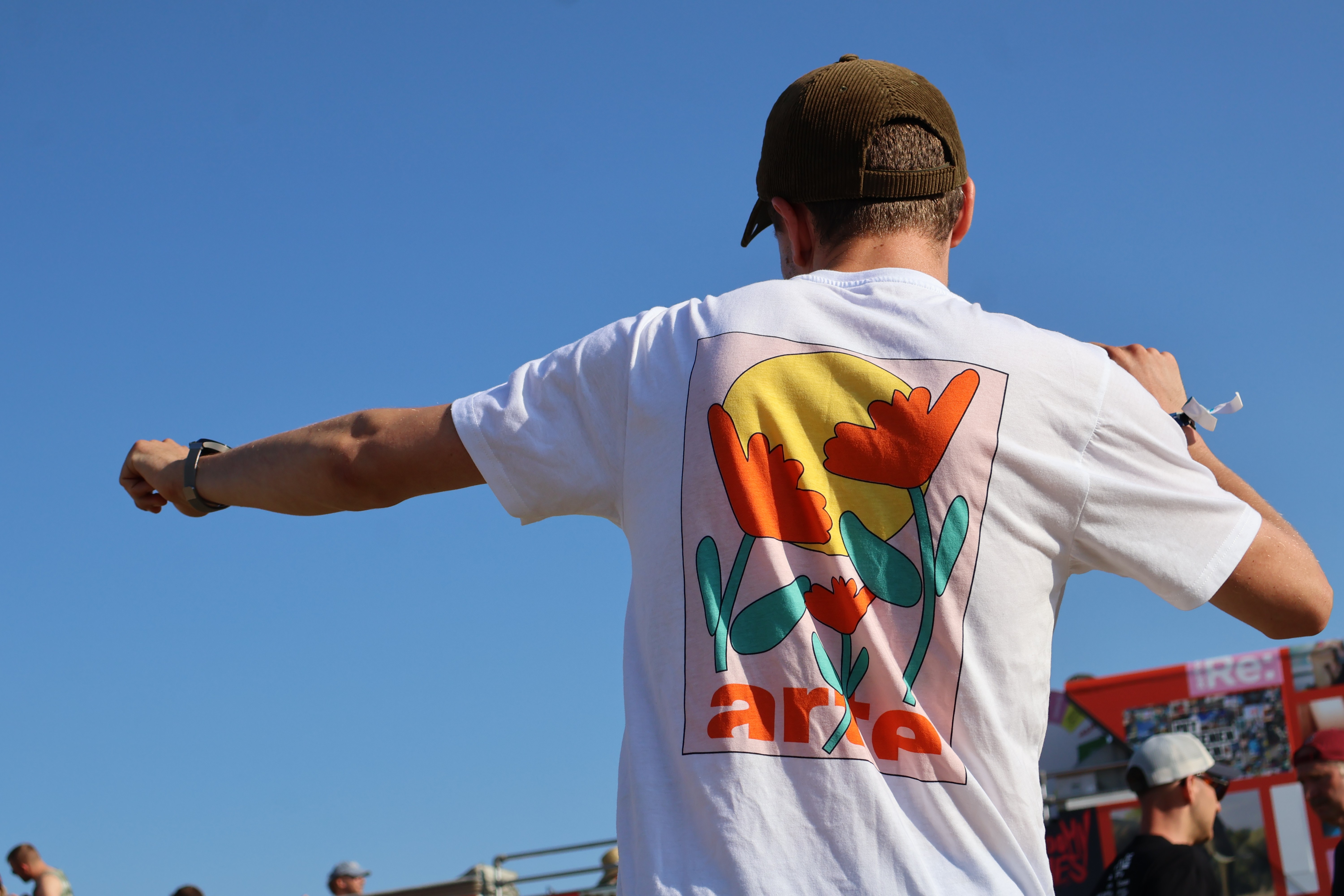 Dancing person, visible from the back, wearing a white shirt with a print in Art Nouveau style showing three orange and green flowers in front of a yellow cercle and the word ‘arte’ written on the bottom, giving summer vibes.