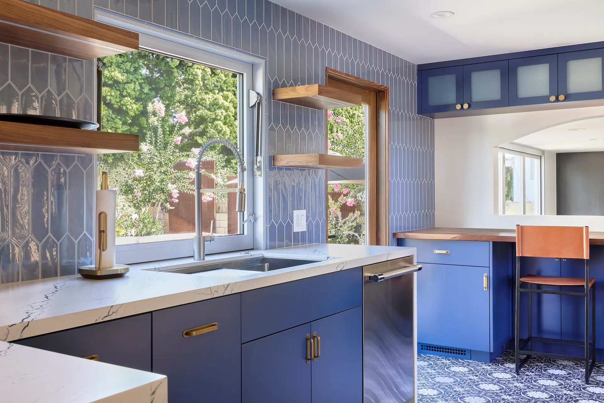 Wide shot at an angle of the kitchen facing the sink wall, featuring an awning window and blue tile backsplash, complemented by brown wooden floating shelves with LED under lighting, in the Huntington Beach Kitchen project. Photo by Todd Huge.