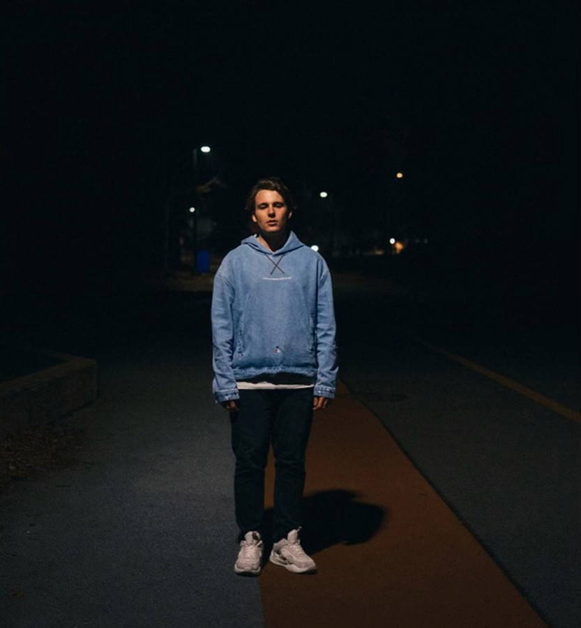 A nighttime photo of music producer Akachi standing on a dimly lit road, wearing a light blue hoodie, dark jeans, and white sneakers.