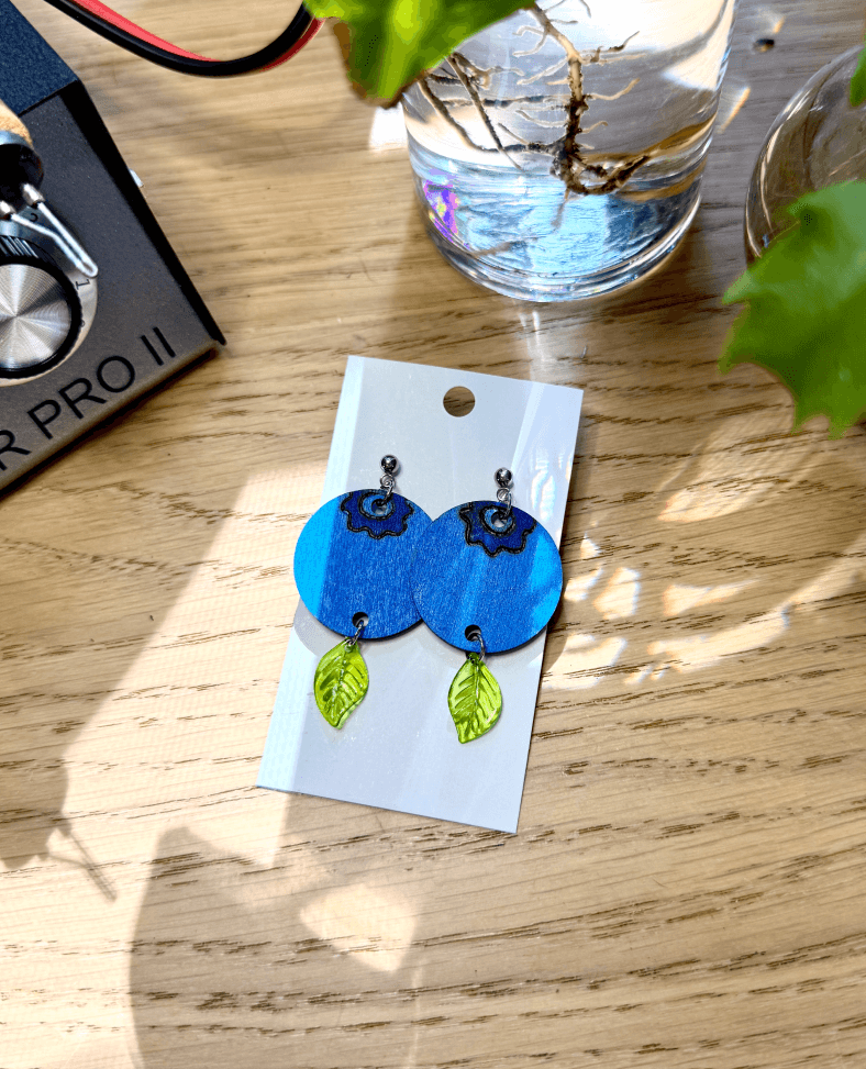 Wood burned earrings depicting large blueberries with dangling leaf charms at the bottom. The earrigns are surrounded by plants, drift wood, and the colwood pro 2 wood burner.