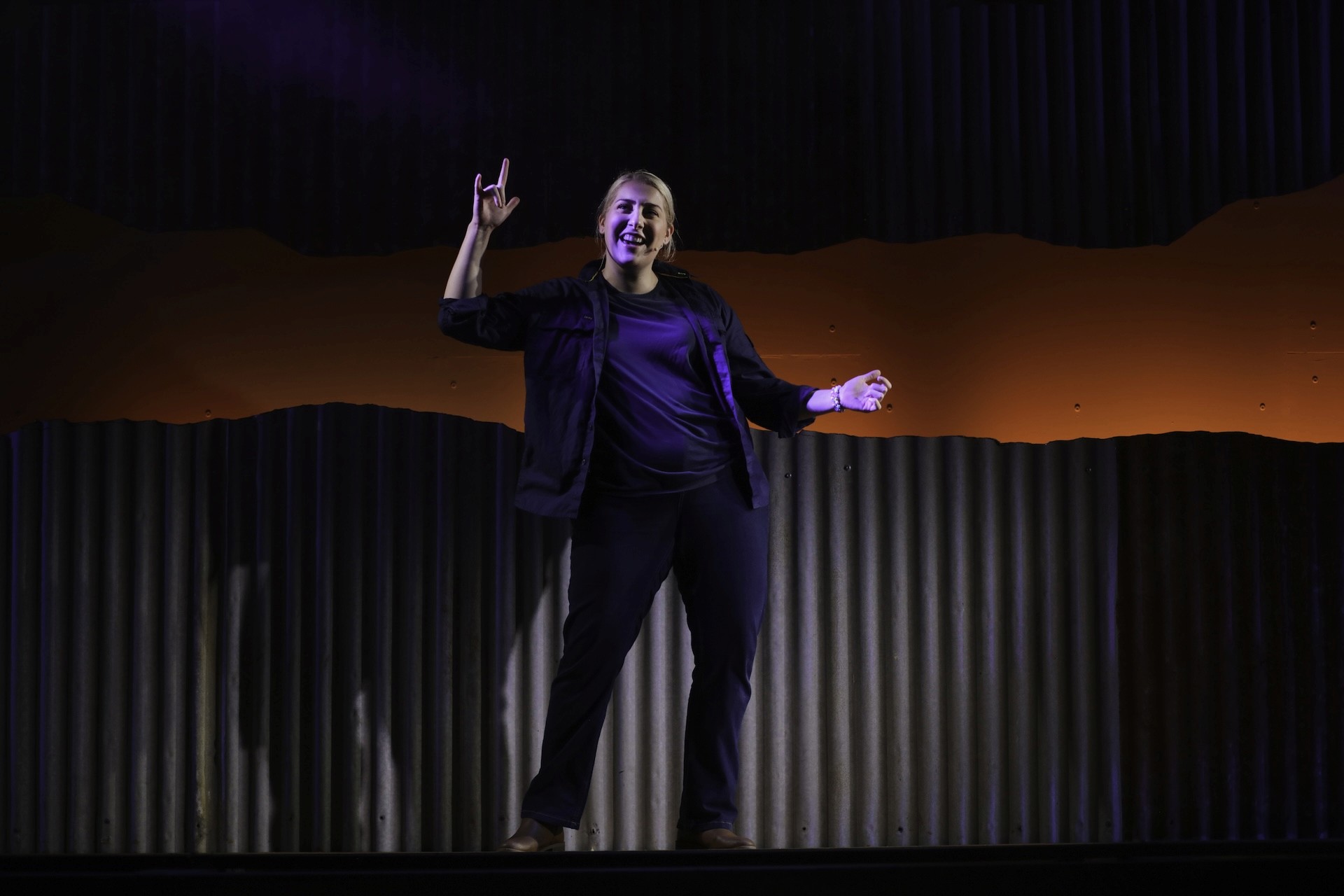 A single performer stands confidently in front of a corrugated metal backdrop washed in orange and purple light. They gesture enthusiastically with one hand raised, delivering a monologue under a focused stage spotlight.