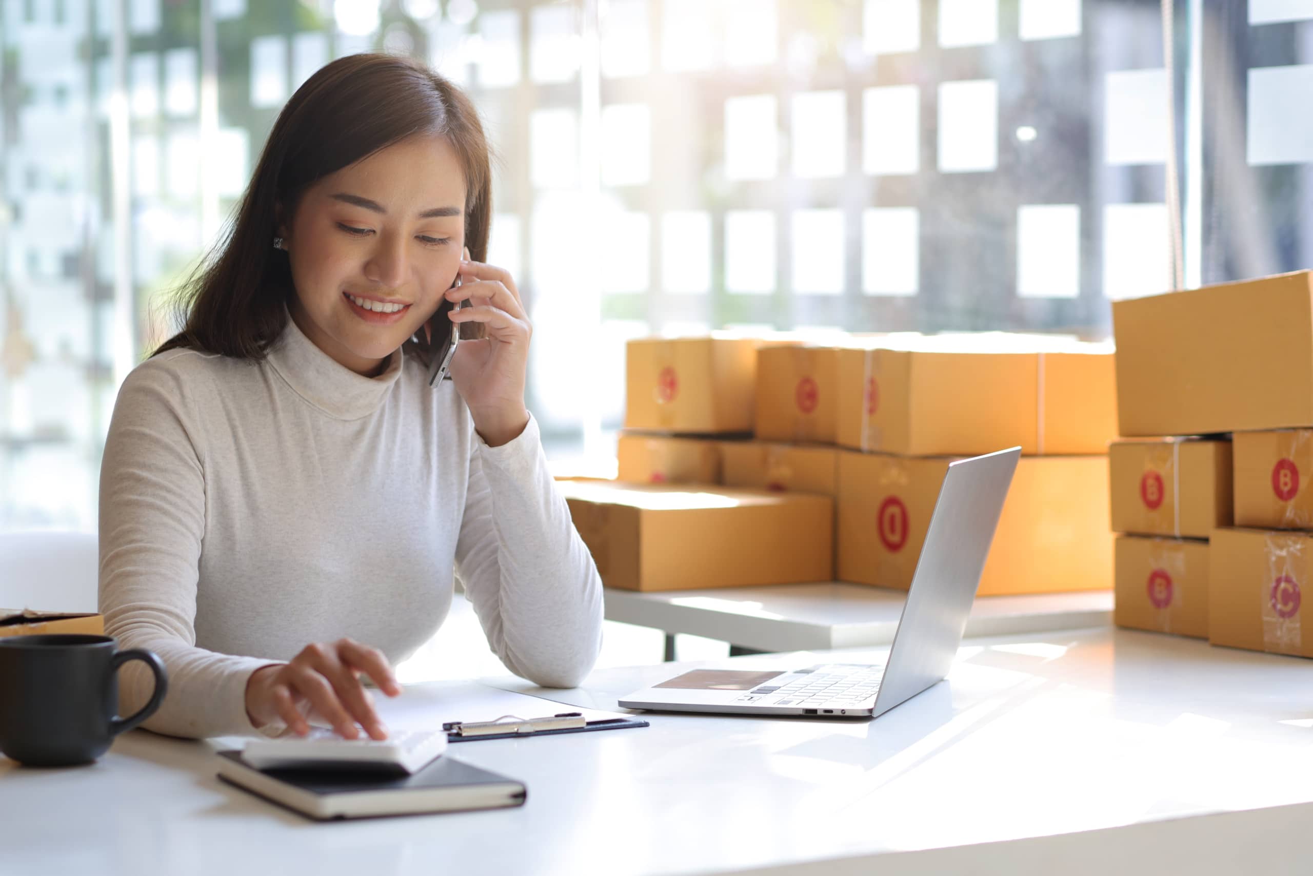 A young woman, e commerce online small start up business owner, calls with a partner and uses a calculator to calculate the cost and shipping costs