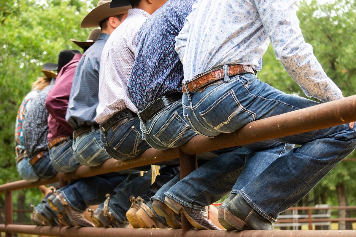 Close-up of cowboys sitting on a wooden fence in denim and cowboy hats during a western-themed event.
