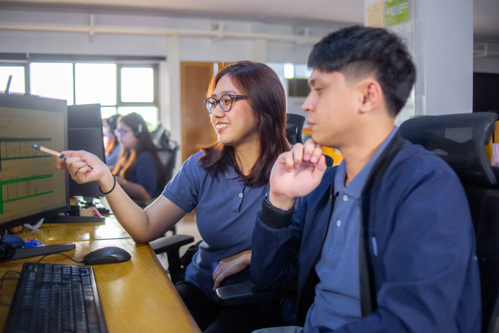 A group of two young professionals collaborate at a shared desk, looking at monitors and discussing their work.