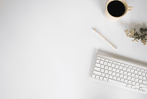 White background with a white and silver Apple Mac keyboard, rainbow pastel patterned pen, cup of black coffee and eucalyptus leaves.