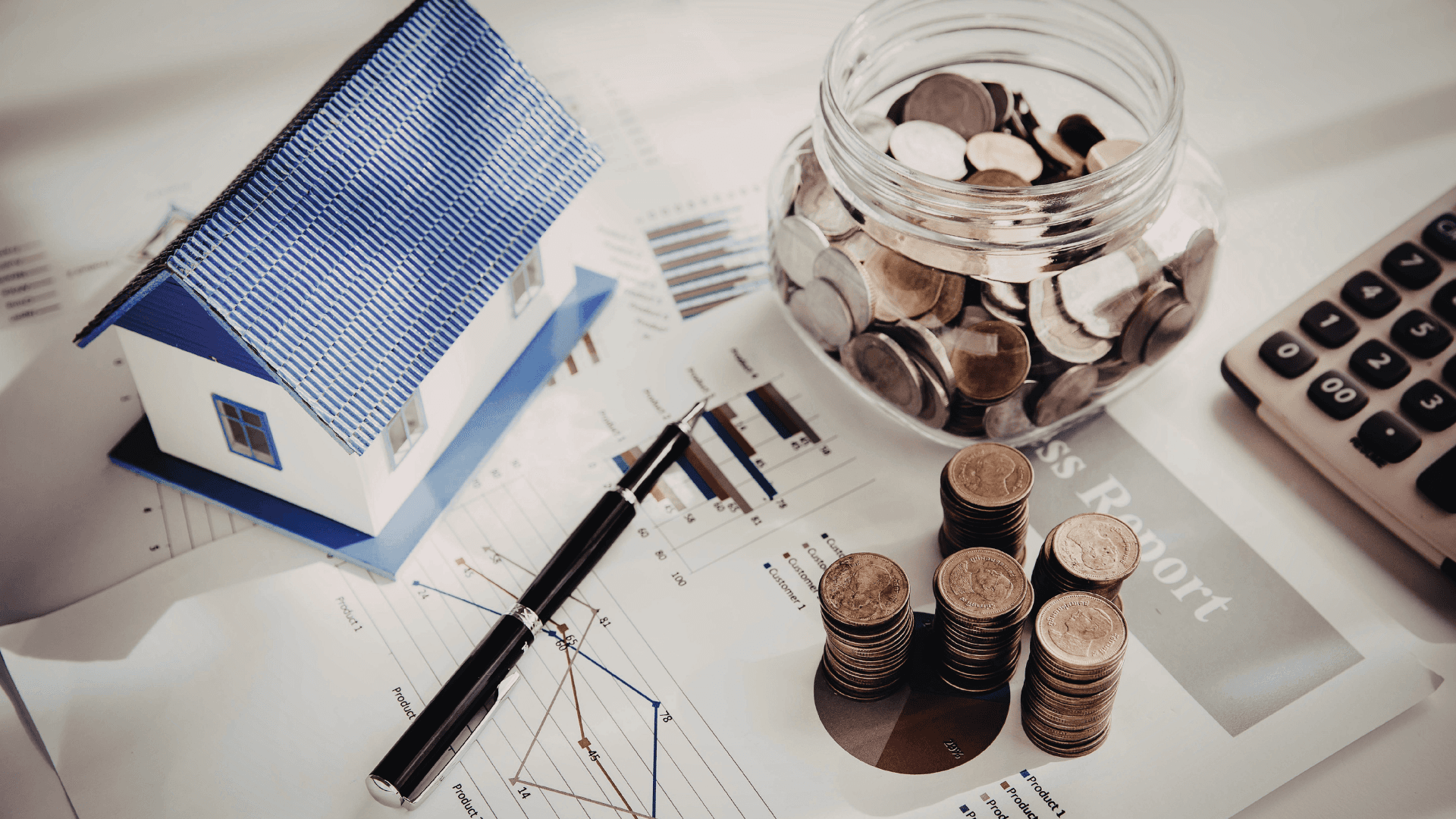 Finanzplanung: Eine Collage aus einem kleinen Hausmodell, einem Glas mit Münzen, einem Taschenrechner und Finanzgrafiken auf einem Schreibtisch.