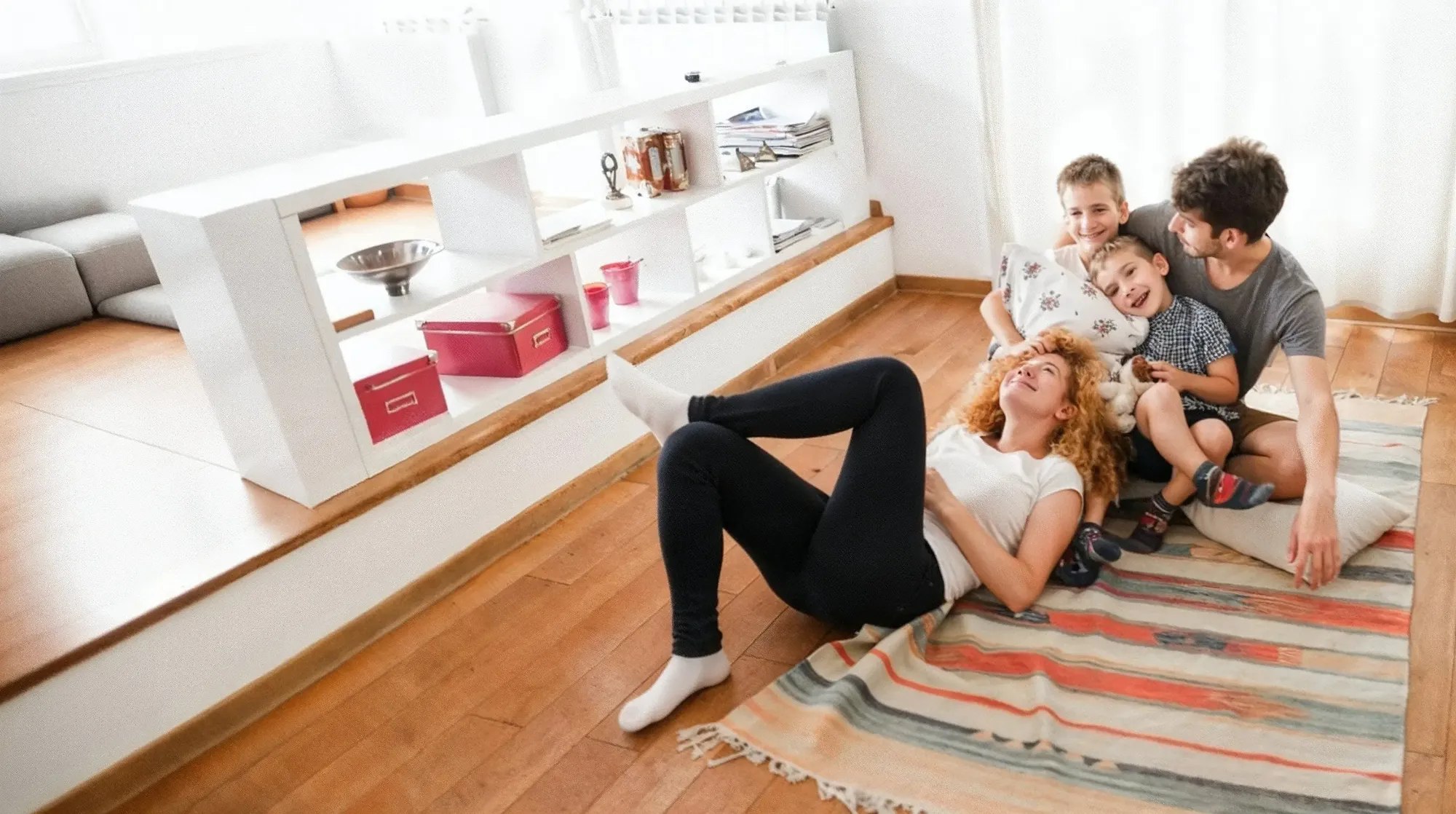 A joyful family of four relaxes together on the floor of their new home, smiling and laughing in a bright living room. The moment captures the comfort, warmth, and happiness that come from finding the perfect home through Chris Lewis Home Loans.