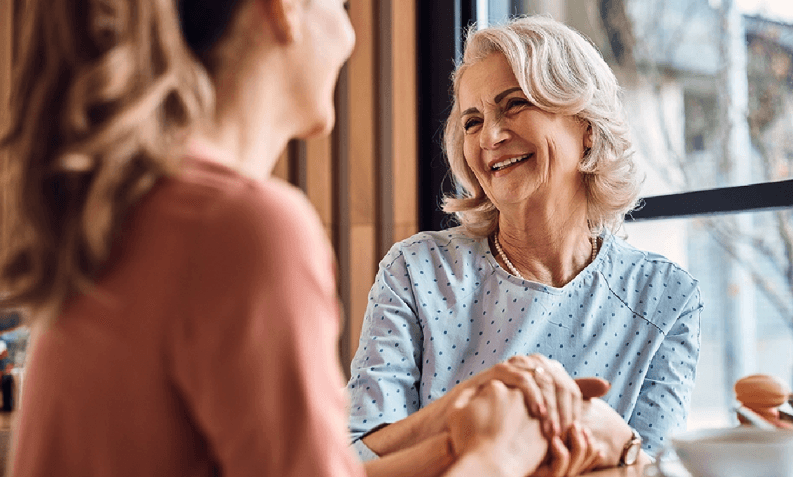 Two women, one younger and one older, share a joyful moment while holding hands and smiling indoors.