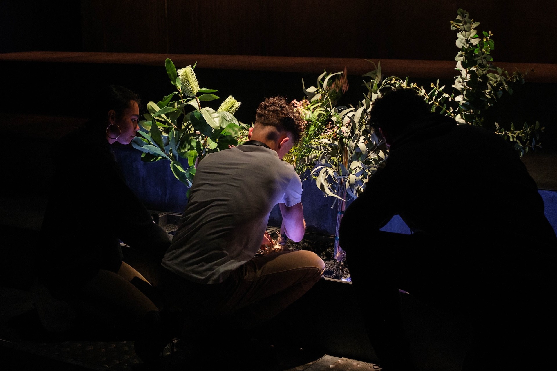 Three young performers kneel in a dimly lit theatre space, tending to a garden bed filled with native Australian plants. Soft blue and amber lighting highlights the foliage as they carefully arrange stems and soil, creating a quiet, reflective atmosphere.