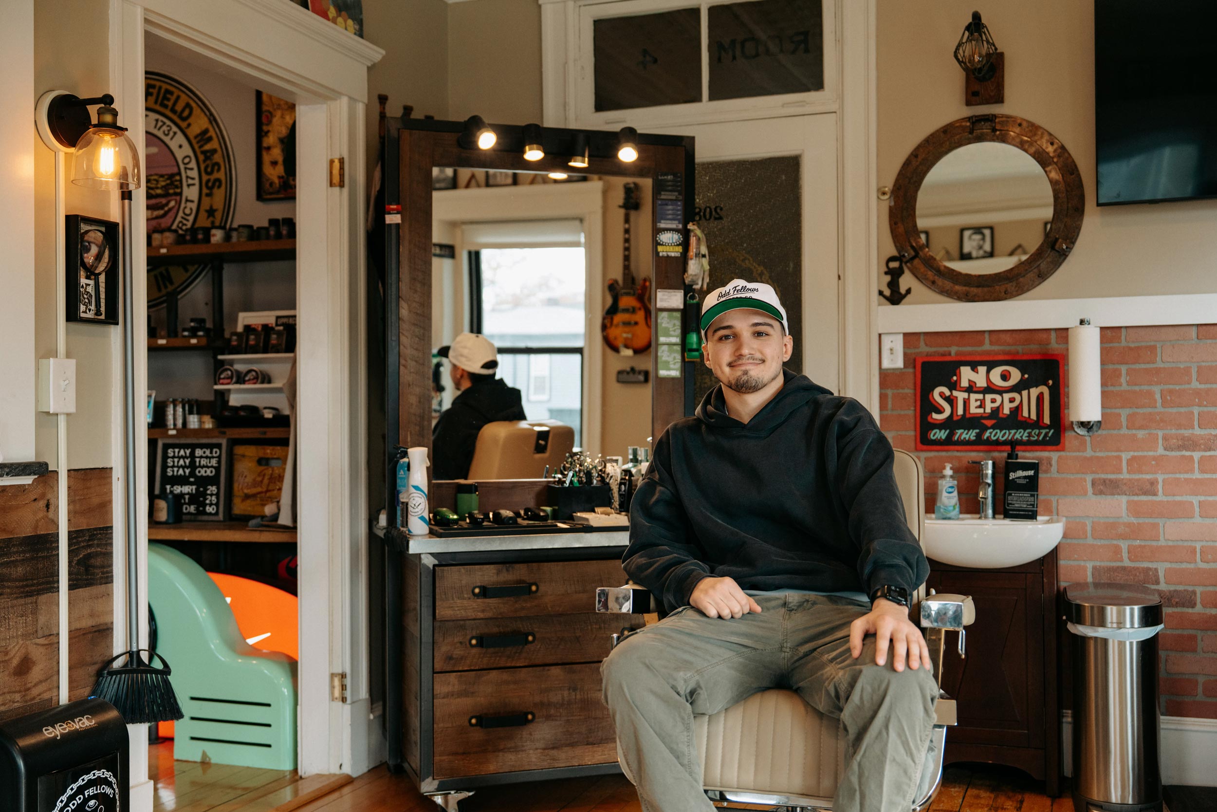 A young man wearing a dark hoodie and a white baseball cap with a green visor sits in a black leather barber chair, looking directly at the camera with a slight smile. He is inside a vintage-style barbershop with a wooden floor, brick wall accents, and a large mirror/cabinet station behind him. A red and black sign on the wall to the right reads, "NO STEPPIN' ON THE FOOTREST!".