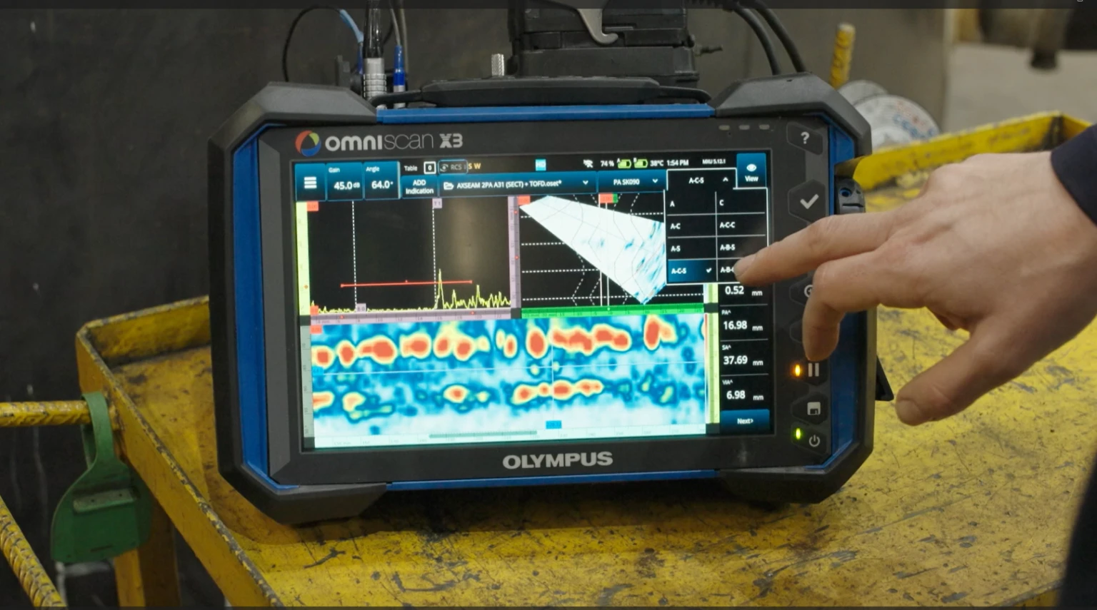 A person points at a tablet displaying a colourful ultrasonic scan with graphs and data analysis, indicating technical inspection in progress.