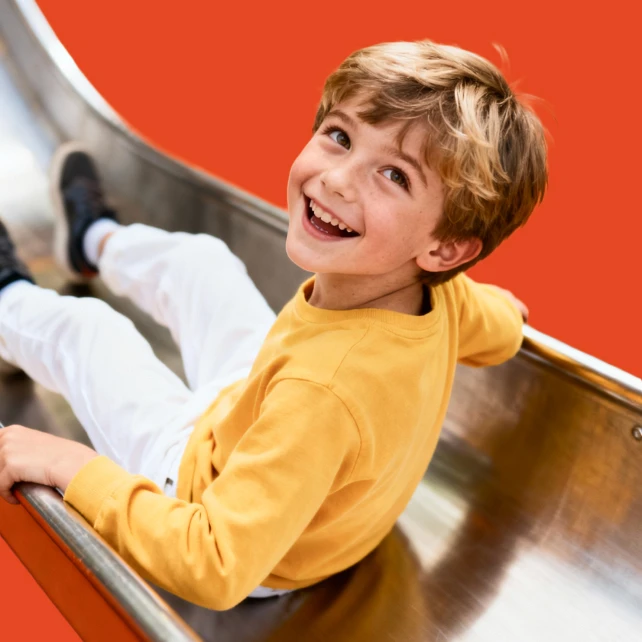A cheerful child with blond hair and a yellow shirt slides down a shiny metal slide against an orange background, smiling brightly at the camera.
