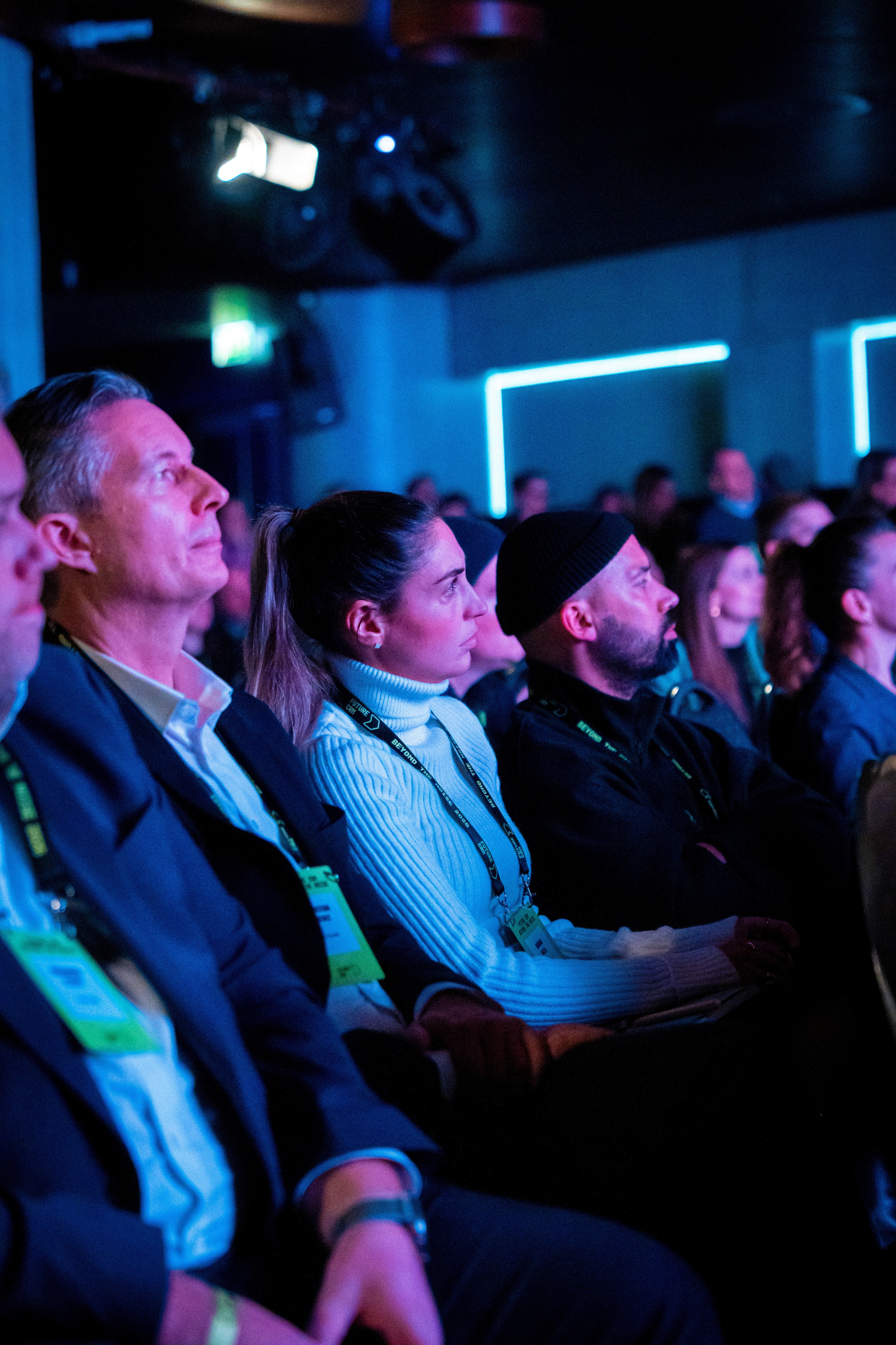 A group of people sit closely together in a dimly lit room, attentively watching something off-camera, wearing lanyards and conference badges, suggesting they are attending a professional event or seminar.