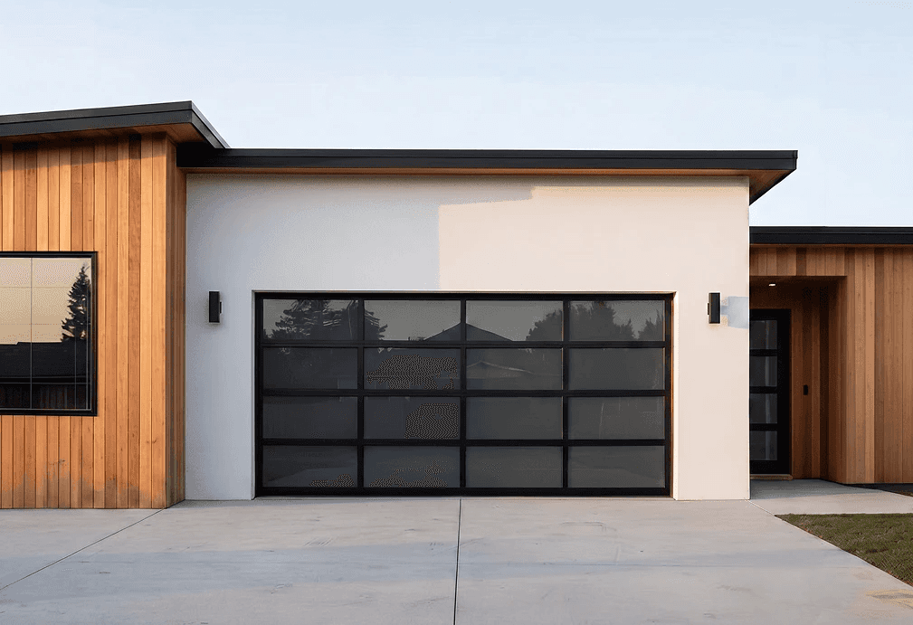 Modern full-view aluminum garage door with tinted glass panels and a black frame. The door is installed on a white minimalist house with natural wood siding and contemporary outdoor lighting.