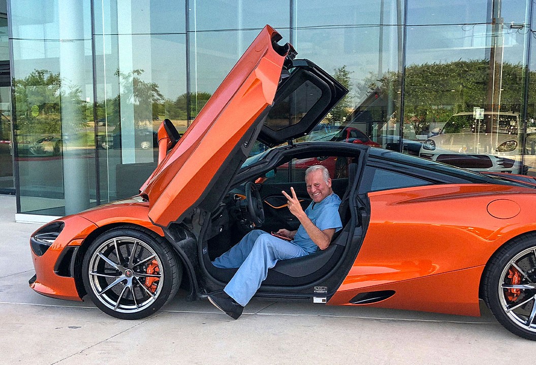 Dr. Billy (Dr. WIlliam Cohn) in blue scrubs sitting in an orange McLaren sports car with its scissor door open, smiling and flashing a peace sign in front of a glass-walled dealership.
