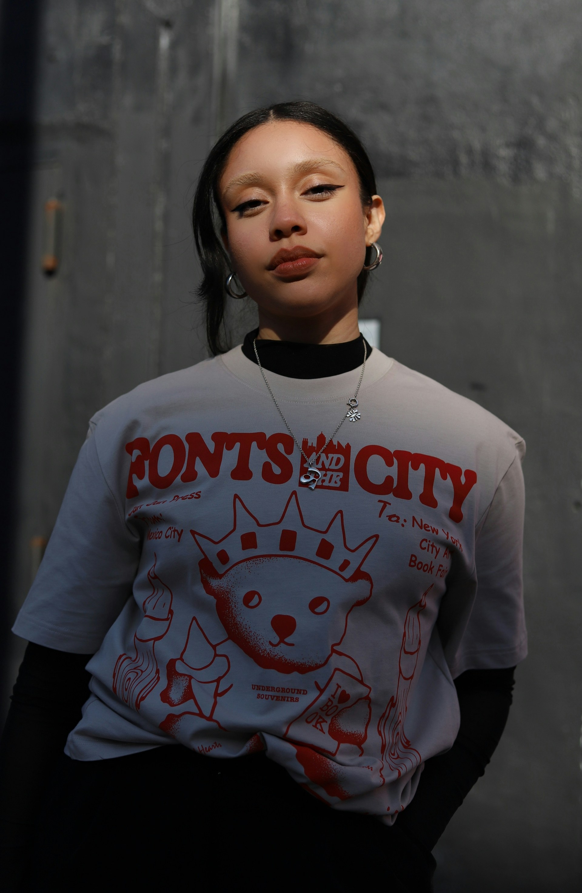 A portrait of a woman with bleached eyebrows and winged eyeliner. She is wearing a grey graphic t-shirt with red text and a bear illustration over a black long-sleeved mesh top and silver hoop earrings.