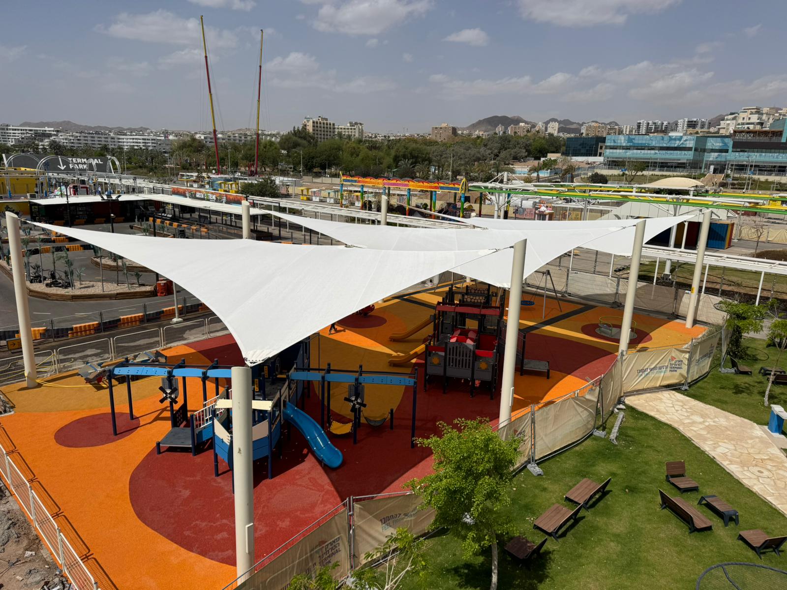 Playground with beige sunshades, slides, and climbing structures on colored surfaces surrounded by benches and green areas.