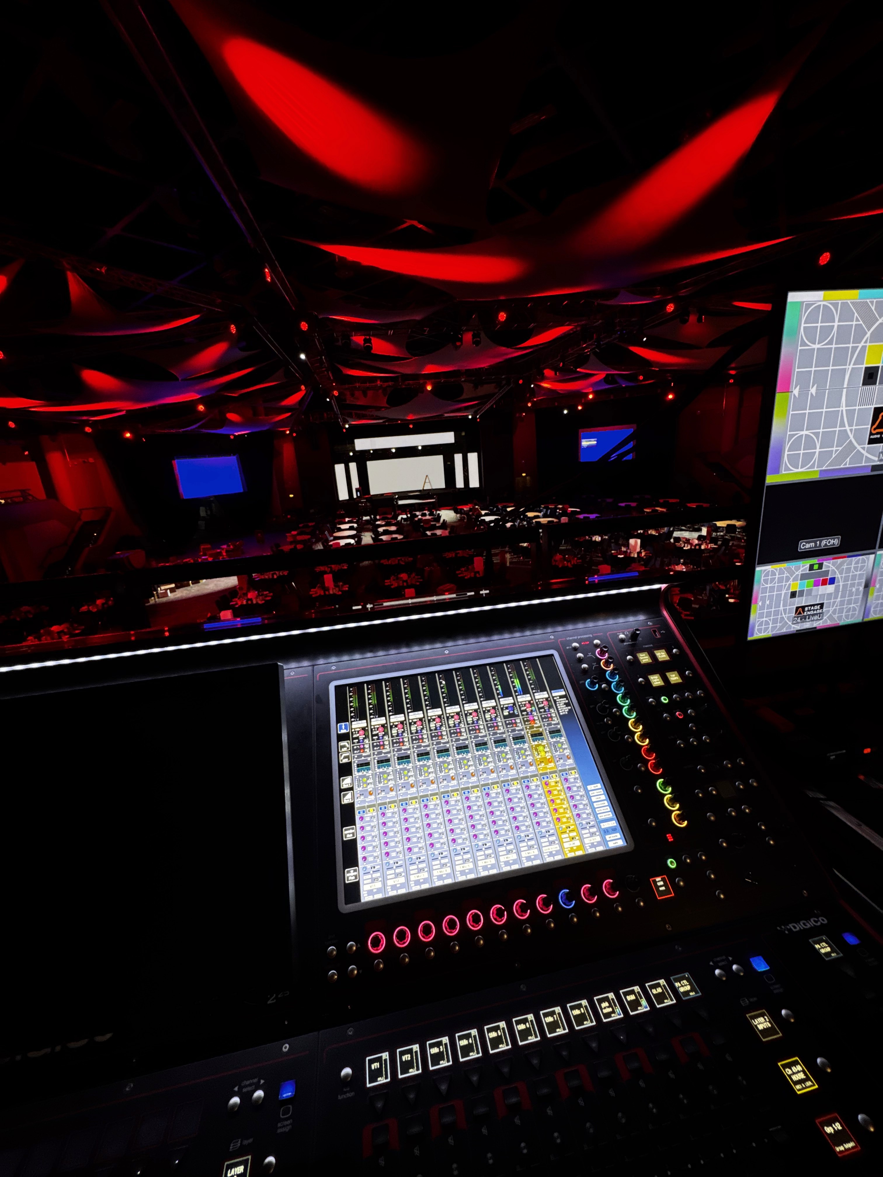 Front of house audio mixing position with digital sound desk overlooking a live event stage, lighting setup and video control screens.