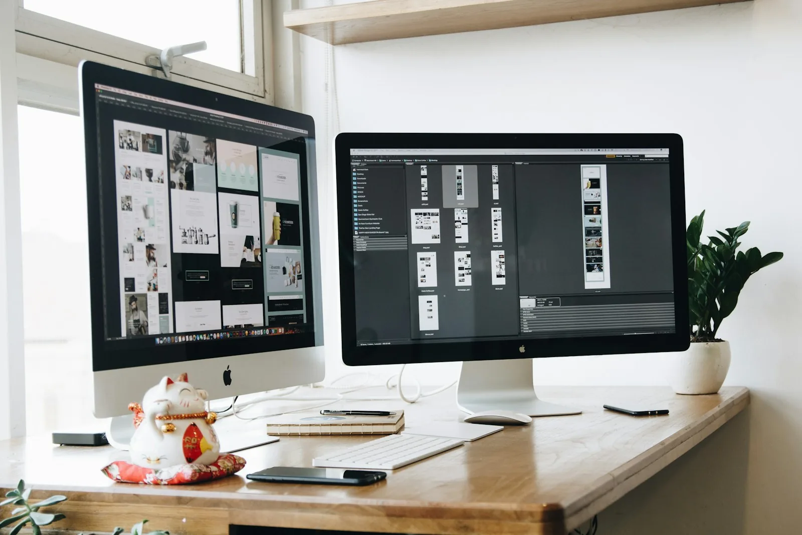 Two desktop monitors on a wooden desk by a window, both showing website page layouts open in a design application