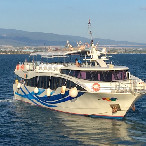 Bílá a modrá výletní loď označená "Lord Byron" proplouvá vodou, s viditelnou krajinou a horami v pozadí.