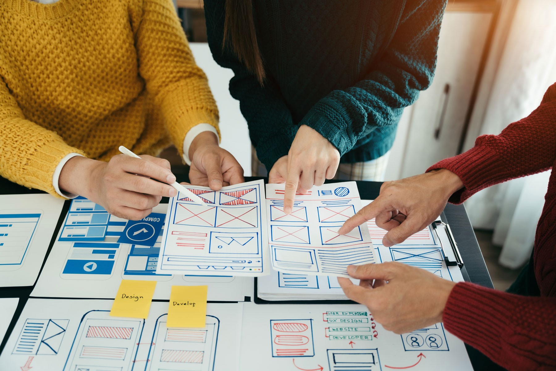 Trzy osoby z zespołu kreatywnego analizujące papierowe makiety i prototypy interfejsu aplikacji mobilnej przy biurku z notatkami