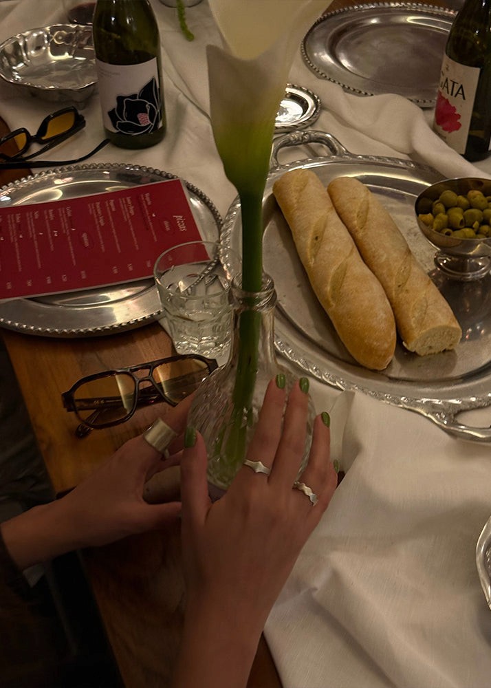 Person holding a white flower in a glass bottle on a table with bread and wine bottles wearing silver rings.