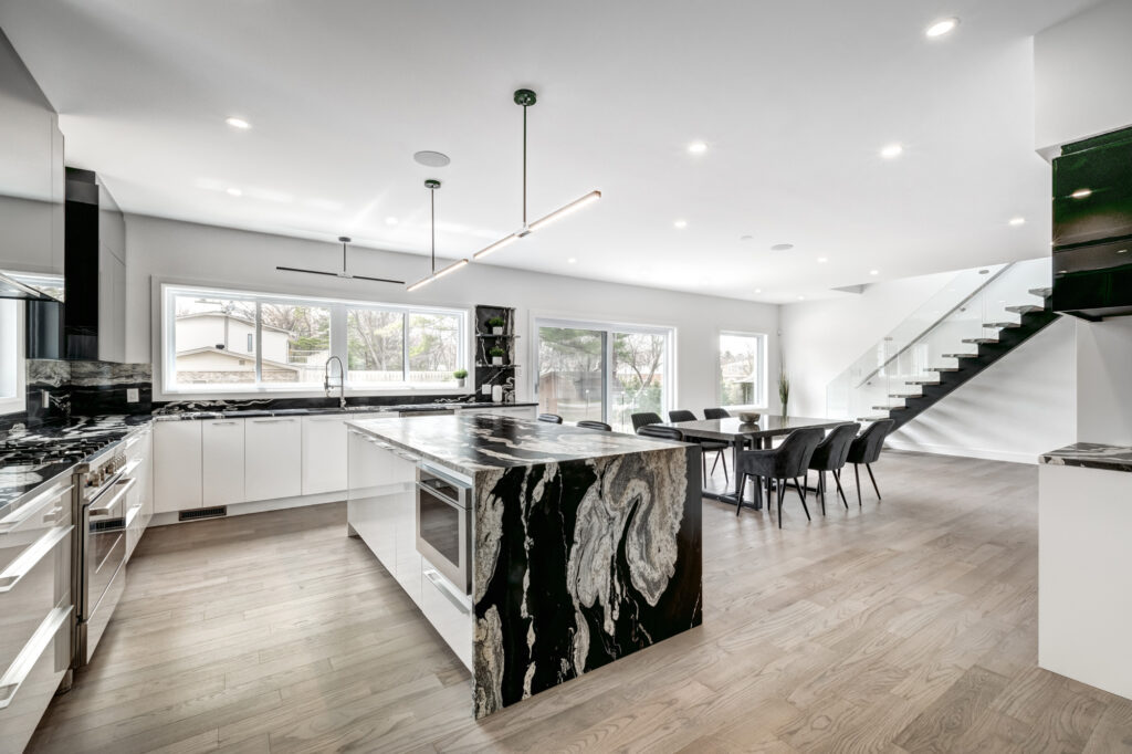 Bright open-concept kitchen and dining area with black and white marble waterfall island, sleek white cabinetry, and glass staircase, showcasing B&Y Reno Corp’s custom kitchen remodeling in a modern luxury home.