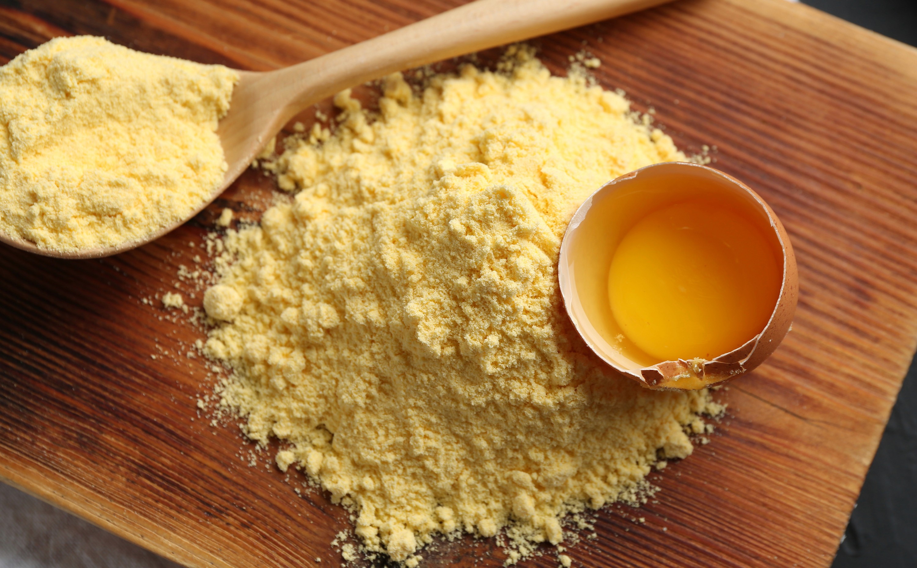 A wooden spoon rests on a pile of vibrant yellow egg powder next to a cracked eggshell holding a rich orange yolk, all on.