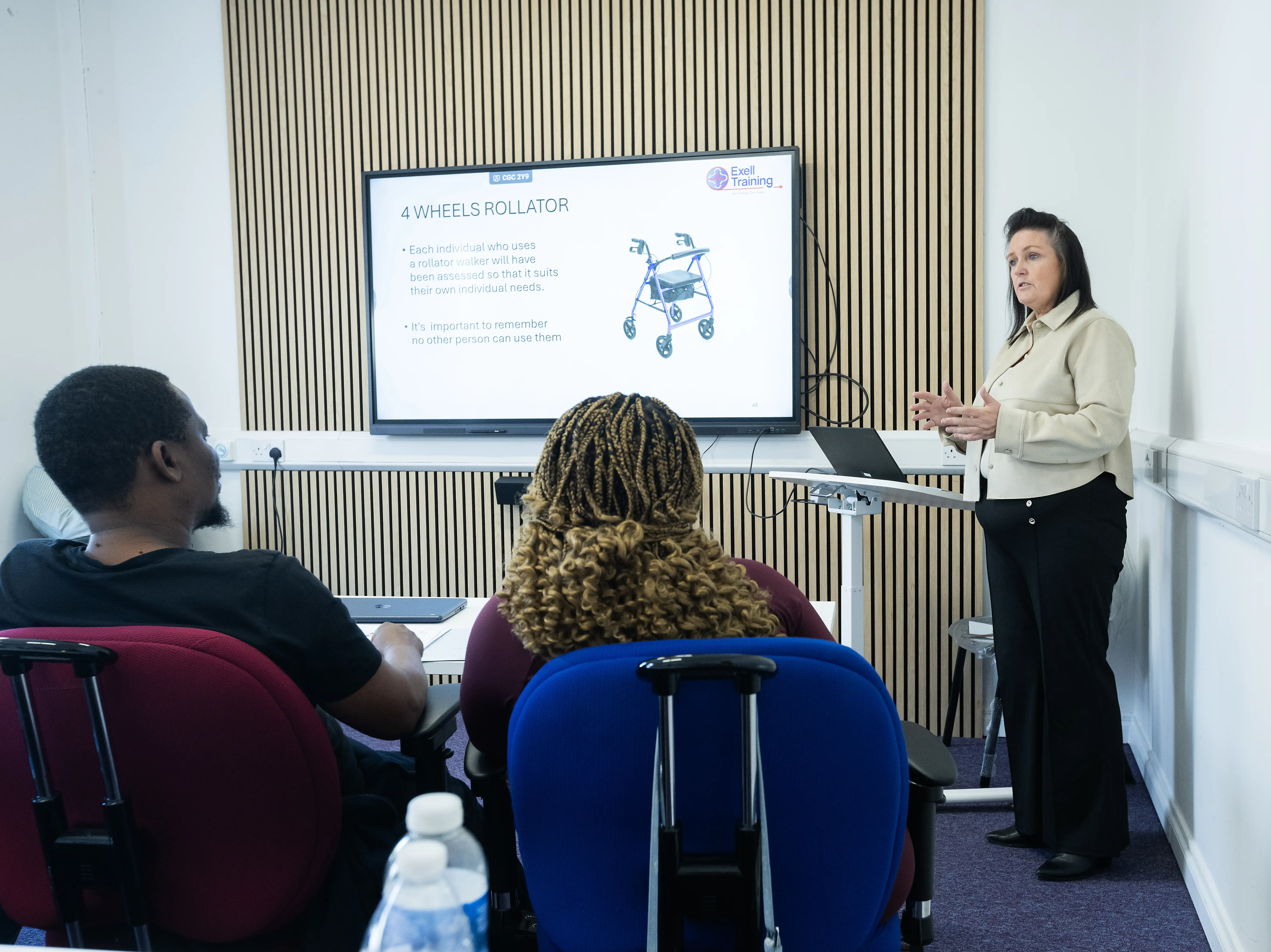 A trainer delivers a mandatory healthcare training session on safe use of mobility aids, specifically a four-wheeled rollator, to a group of adult learners at Exell Training. A presentation slide is displayed on a screen behind her.