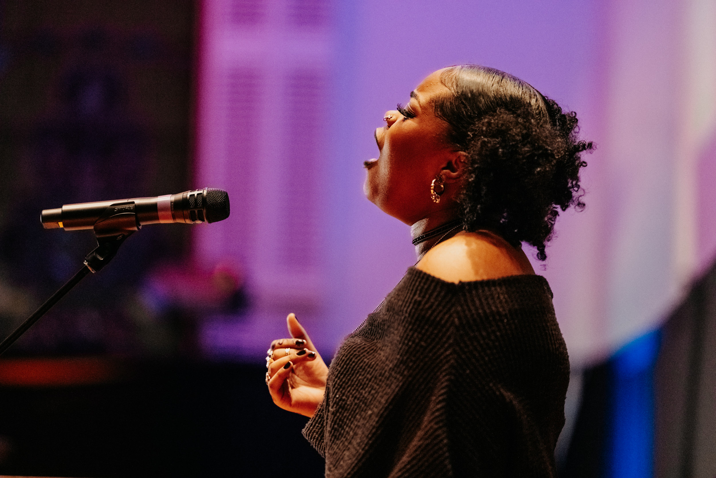 Performer singing into a microphone at the Queer Open Mic (QOM) hosted by be; community in Bridgewater, Massachusetts