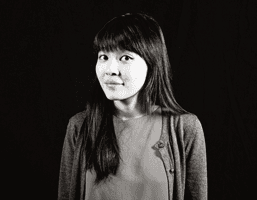 A black and white portrait of a young woman with long, dark hair and bangs. She wears a light-colored top and a cardigan or sweater over it. The background is uniformly dark.