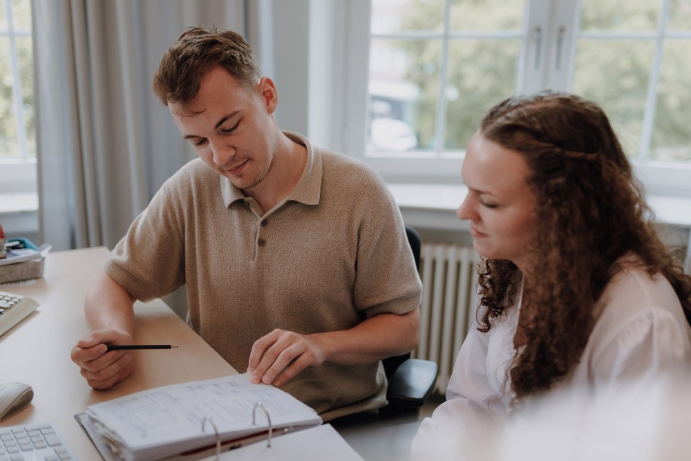 Zwei Personen sitzen nebeneinander am Tisch und gehen gemeinsam Dokumente durch.