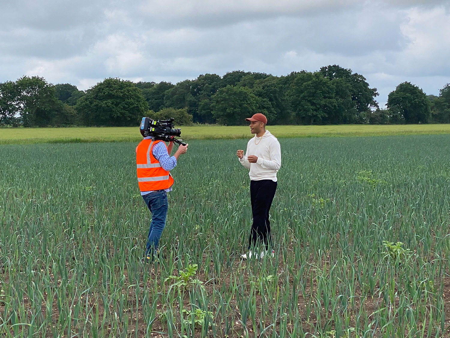 Joe standing in a field delivering a piece to camera. There is a camera operator wearing an ornage hi-vis vest.