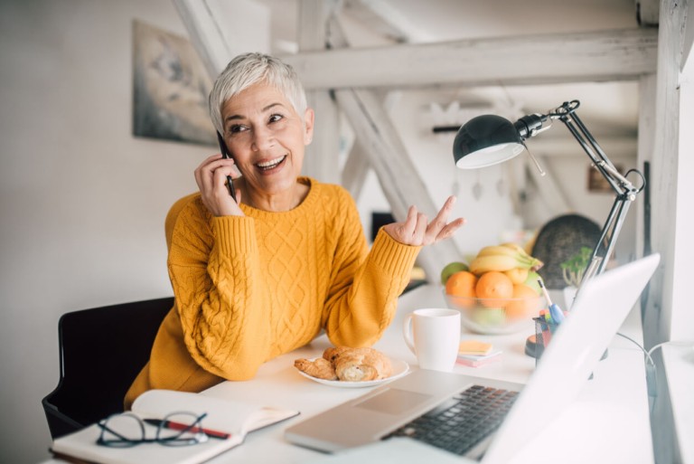 Woman working from home, talking on the phone while sitting at a desk with a laptop and coffee.