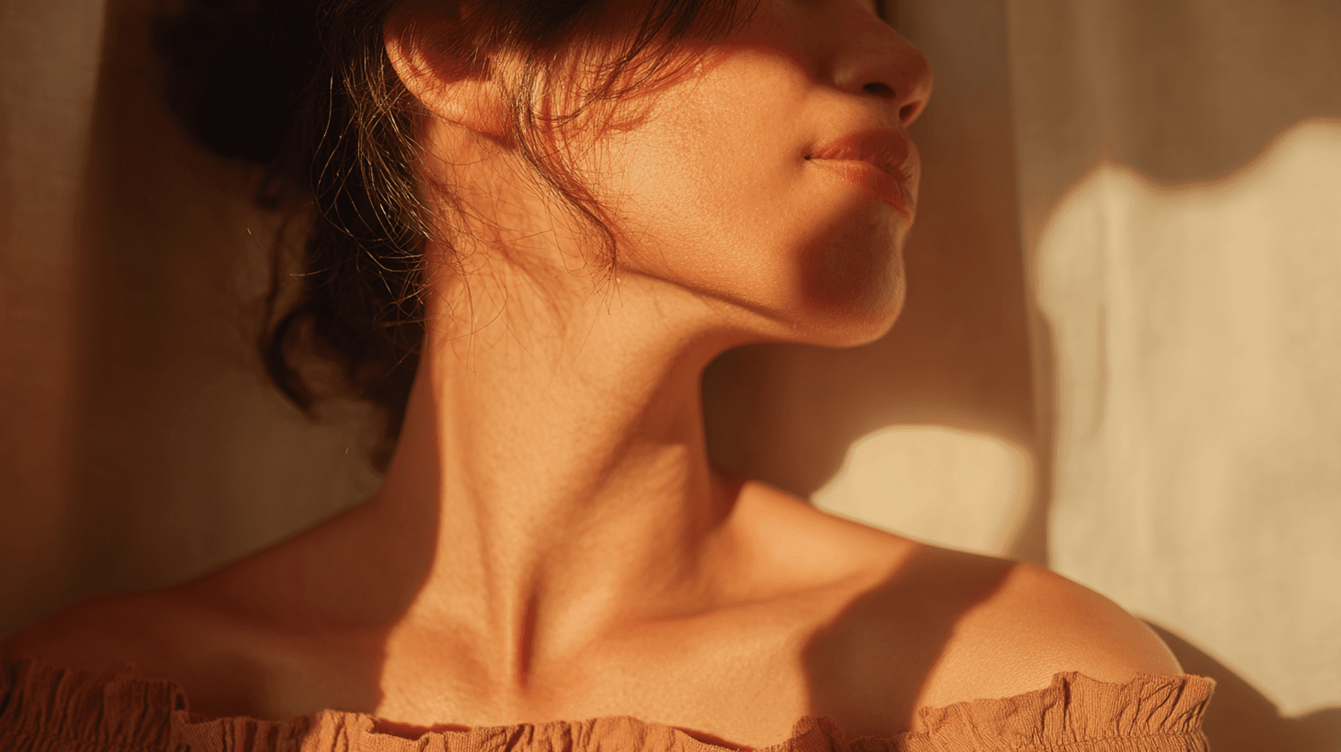 Close-up of a person’s neck and collarbone in warm sunlight, with soft window shadows creating a calm, intimate mood.