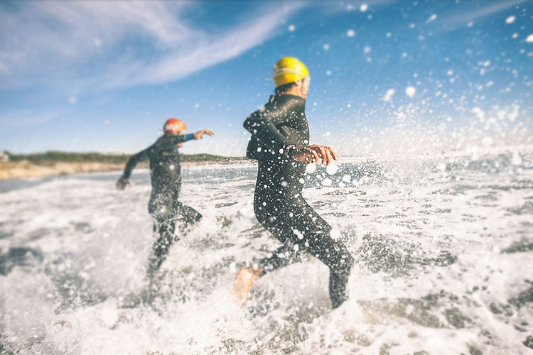 Two people wearing wetsuits are running through shallow water. The person in the foreground wears a yellow swim cap and is running away from the camera. The person further back wears an orange swim cap and is running in the opposite direction. The water is splashing around them, and a sandy beach is visible in the background under a blue sky with scattered clouds.
