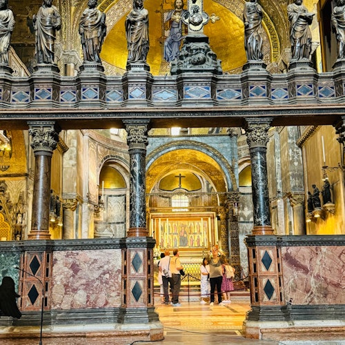 Interior of an ornate church with statues, arches, and people observing a golden altar under intricate decorations.