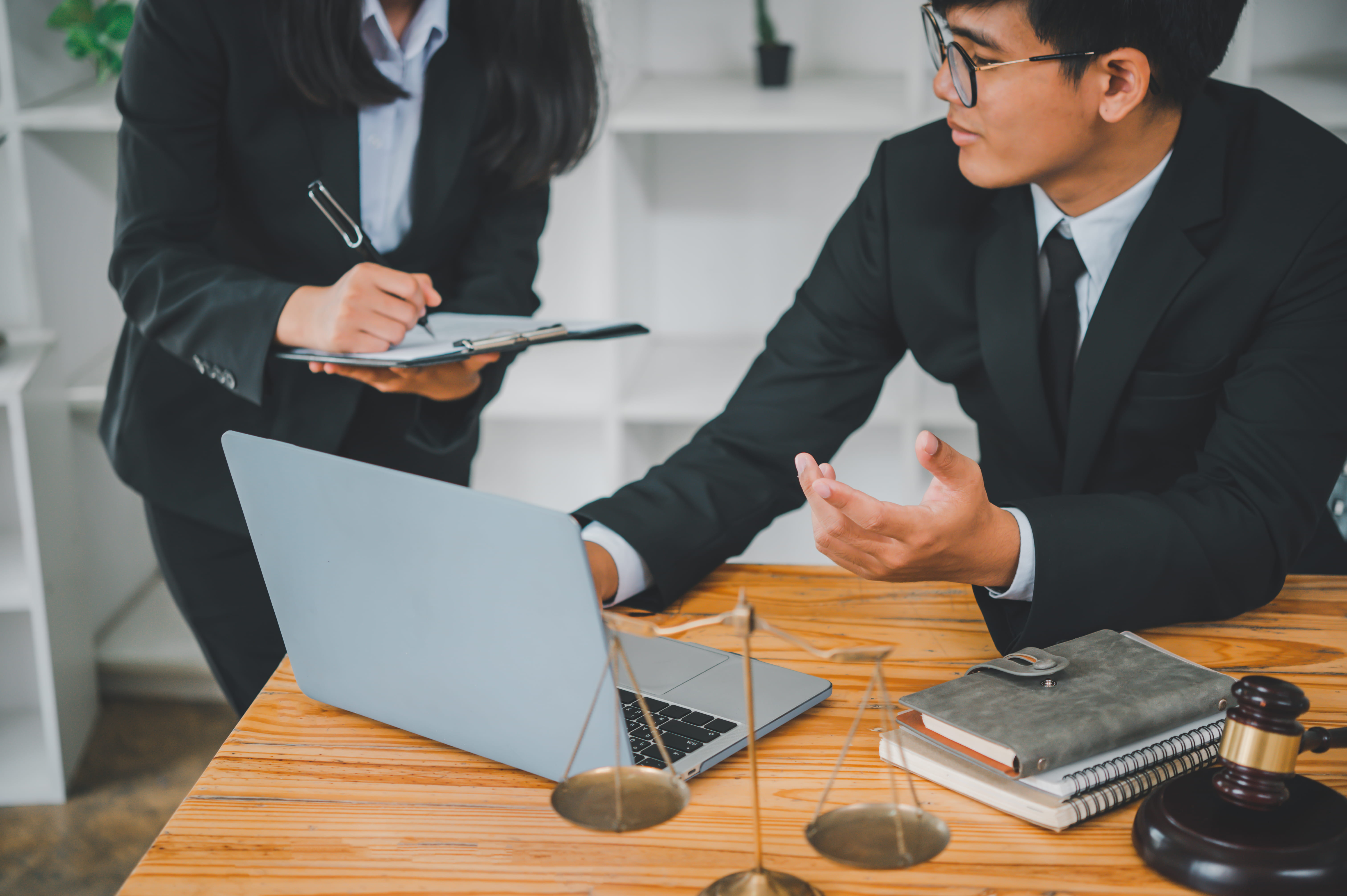 Two legal professionals in suits discuss work at a desk with a laptop, clipboard, scales of justice, notebooks, and a judge’s gavel.