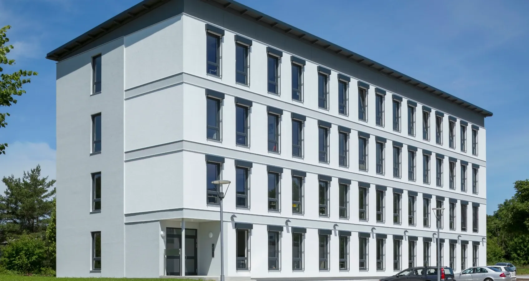 Modernes, mehrstöckiges Gebäude mit weißer Fassade, grauem Dach und vielen Fenstern unter blauem Himmel.